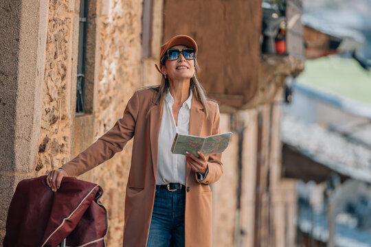 Smiling Caucasian woman tourist wearing sunglasses and a cap, holding a map while exploring a narrow old town street, travel and adventure concept