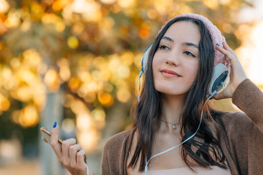 young woman smiling and listening to music with headphones, holding a smartphone, autumn park background with golden bokeh leaves