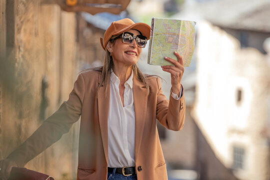 Smiling mature woman tourist reading a city map outdoors, enjoying a sunny travel adventure in a European old town