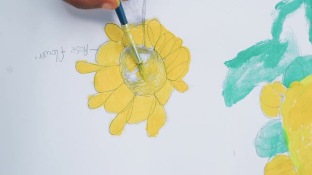Hand paints yellow details on sunflower sketch gradually filling in center and petals. white paper background shows other partially completed colorful drawings with handwritten labels