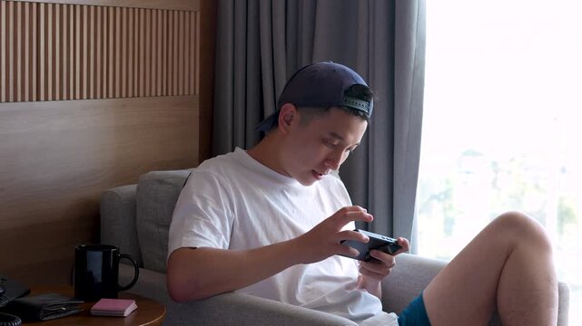 shot of a happy young Asian man in a backward cap relaxing on an armchair, focusing on his phone and play games 