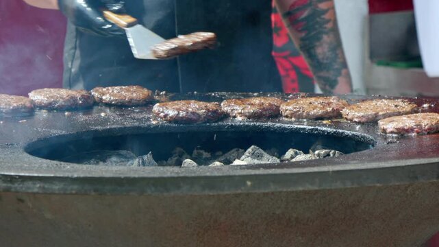 Charcoal grill steams as meat sizzles. Hands flip patties with metal spatulas. Glowing coals heat the grilling surface. Cooked meat releases fragrant smoke. Crisp, smoky burgers sizzle on grill