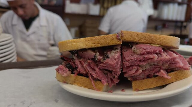 Close-up of a classic pastrami on rye sandwich, packed with sliced cured meat and served on a plate, with busy deli workers preparing food in the blurred background