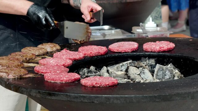 Cook places burgers on hot griddle over glowing coals. Hands skillfully turn patty as sizzle fills air. Smoke rises gently from charcoal beneath patties. Busy cook prepares food with care and speed