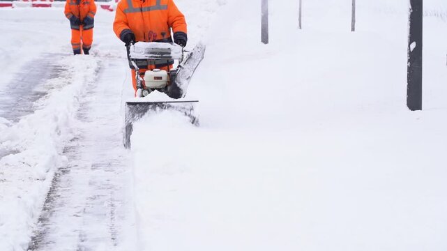 Worker clears snow with heavy machinery. Another worker watches nearby. Snow blankets street and poles. Scene shows winter clearing effort. Ideal for urban winter maintenance projects