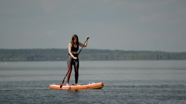 Woman stands on bright paddleboard. She rows gently across calm lake surface. Distant green trees frame hazy horizon. She appears relaxed, enjoying tranquil water