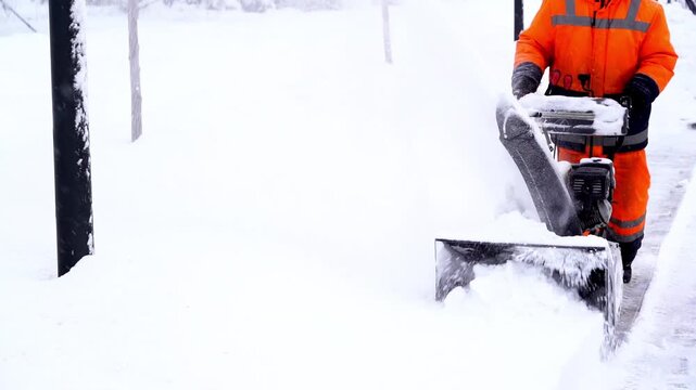 Worker clears snow with machine. Snow falls gently around him. Bright orange suit stands out in white. He moves steadily through the path. Snow flies as machine grinds ahead