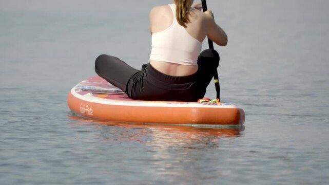 Woman sits cross-legged on paddleboard. She paddles gently through calm water. Back view shows focused, peaceful demeanor. Ideal for wellness, fitness, or nature themes. No animals or plants present