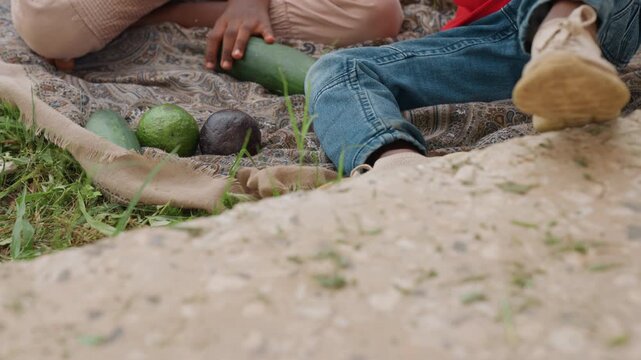 Two children sit together outdoors. One child wears blue jeans and other beige pants on patterned blanket with avocados cucumber cloth grass visible legs