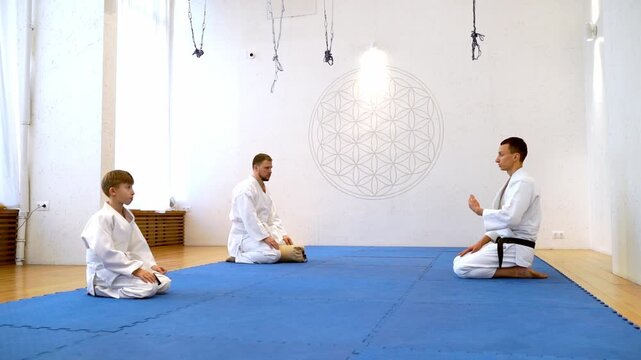 Three martial artists sit on a blue mat. Man with a black belt speaks to two younger students. They are in a quiet, brightly lit dojo. The setting includes a large geometric wall design