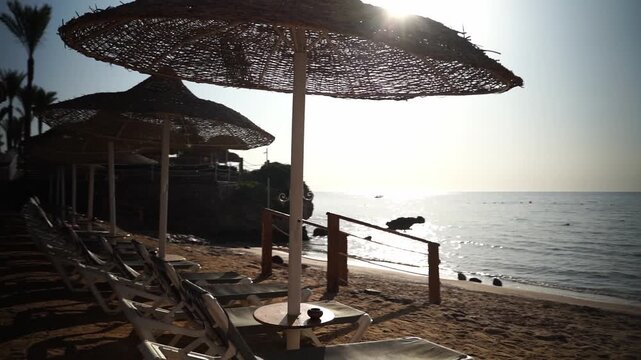 Sun sets over calm sea, casting golden glow. Beachside loungers and thatched umbrellas line the shore. Rocks and gentle waves frame the tranquil scene. No people present, peaceful moment embraced