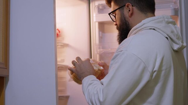 Man removes packaged food from an open refrigerator and checks it, illustrating food storage, freshness, household routine and product inspection.