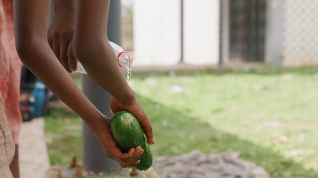 Hands holding and washing fresh cucumbers under running tap with clear water splashing. Hands manipulate vegetable while pouring from plastic bottle in outdoor environment