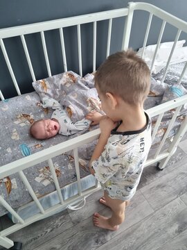 Vertical high angle full shot of a toddler boy looking at a crying newborn baby sibling lying inside a white wooden crib, grey pattern bedding, natural morning indoor lighting, cozy home room