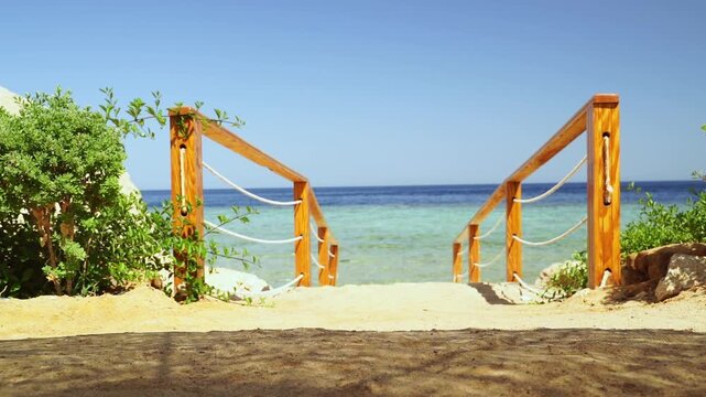 Wooden stairs lead to turquoise sea. Sandy path curves gently toward ocean. Bright blue sky frames tranquil beach scene. Natural greenery borders the entrance. Perfect for vacation, travel