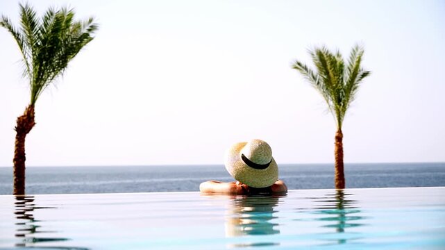 Woman relaxes in infinity pool beside palm trees. Sunlight glows softly over calm ocean water. She rests, serene, with hat on head and eyes closed. Perfect for vacation, luxury
