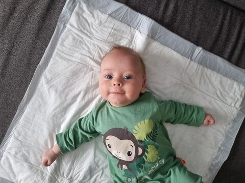 High angle full shot of a cute happy baby lying on back on a white blanket looking at camera, infant wearing a green bodysuit with a monkey print, soft natural daytime indoor lighting