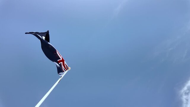 Slow motion and low-angle view looking up at the Australian national flag fluttering on a white flagpole against a vast, clear blue sky. Negative space for design. National pride, patriotic symbolism.