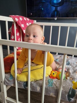 Vertical home photo of a real baby boy looking away through the white wooden bars of a crib wearing yellow long sleeve clothing soft indoor daytime light background