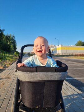 Vertical outdoor photo of a happy smiling real baby boy sitting in a black stroller bassinet on a paved path wearing light blue bodysuit bright sunny summer evening daylight clear blue sky background
