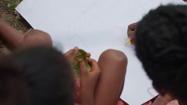 Two children sit together outdoors. One child draws design using pencil while other holds green object. White sheet of paper with sketch visible on ground
