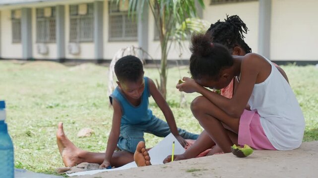 Children sit outdoors on grass near building. One child draws with pencil and paper while another plays nearby. third person is partially visible in background