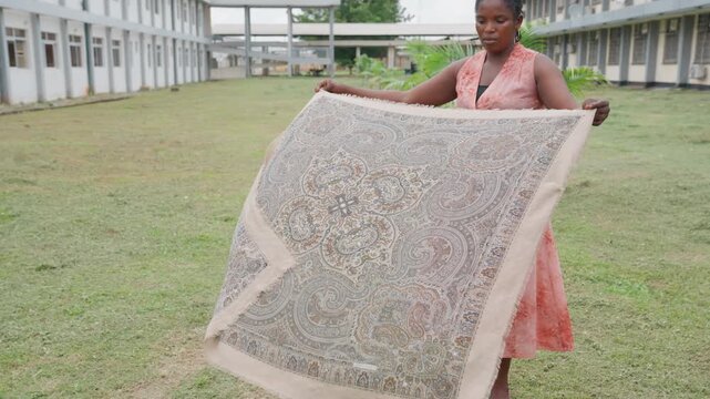 Lady in pink dress displays ornate paisley patterned cloth on grassy ground with building background. scene is steady showing full view of her holding large textile outdoors