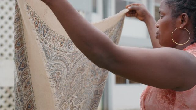 Woman examines patterned fabric outdoors. Woman holds textile up for closer look assessing its design details against architectural backdrop with lattice and windows