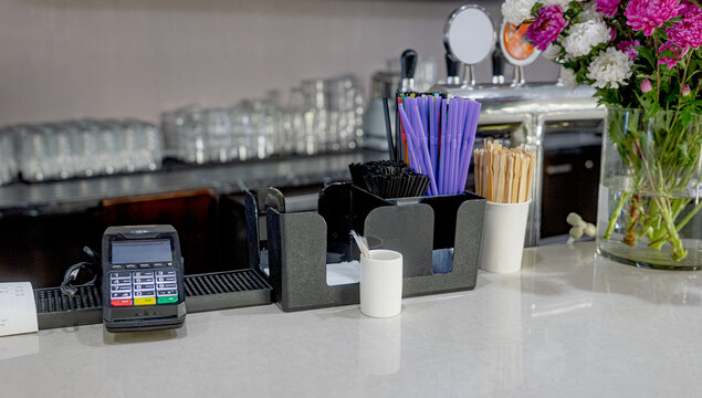 Cafe service counter featuring woven baskets with sugar packets, wooden stirrers in a cup, bottled water and a coffee machine behind the bar
