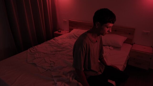 Anxious man sitting alone on the edge of his bed, lost in worried thoughts and emotional isolation, portraying depression, loneliness and psychological distress in a dark low key red bedroom