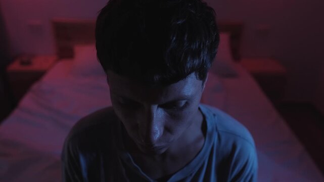 Man sitting on his bed overwhelmed by racing thoughts and uncertainty, struggling with decision paralysis, overthinking and distress in a dark bedroom with blue and red low key lighting
