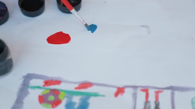 Close up view of white surface with red blue paints and partially completed colorful drawing. brush applies small amounts of paint to paper while various colors are visible in containers