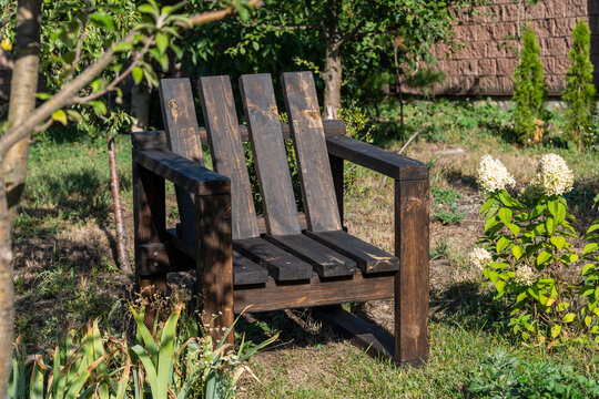 Handmade dark wooden armchair in sunny green garden. Rustic wood adirondack chair on backyard lawn. Comfortable outdoor furniture set in summer garden with blooming white hydrangea flowers