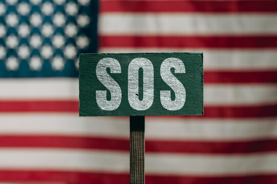 Green SOS sign with American flag background. Plaque with white handwritten SOS letters, closeup. Defocused USA flag background for political crisis, social distress or economic emergency concept