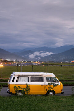 Yellow vintage camper van stands beside illuminated Batumi airport runway under evening sky. Roadside location near security fence attracts plane spotters watching aircraft takeoffs and landings.