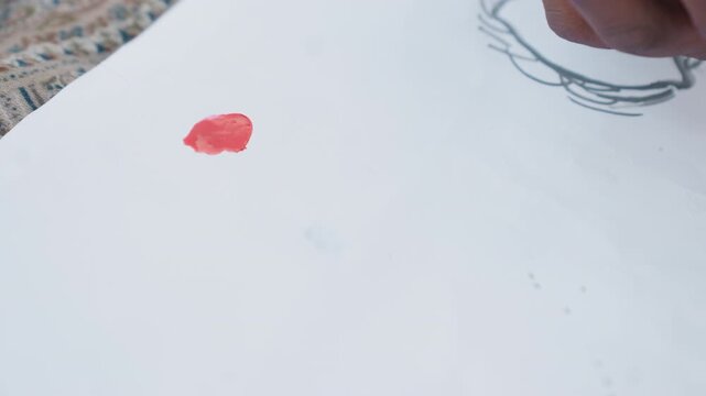 Red paint is carefully added in small dollops onto blank sheet part of ongoing artwork featuring initial pencil sketches. scene focuses on texture and placement of each red mark