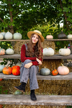 Young woman in straw hat sitting among colorful pumpkins at an autumn harvest display, rustic farm scene with hay bales and seasonal decorations.