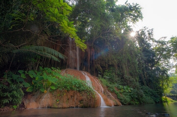 A waterfall cascades down from a limestone cliff, with a river in the forest in front, bathed in sunlight. © Pongsorn