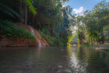 A waterfall cascades down from a limestone cliff, with a river in the forest in front, bathed in sunlight. © Pongsorn