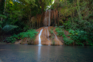 A waterfall cascades down from a limestone cliff, with a river in the forest in front, bathed in sunlight. © Pongsorn