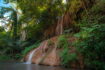 A waterfall cascades down from a limestone cliff, with a river in the forest in front, bathed in sunlight. © Pongsorn