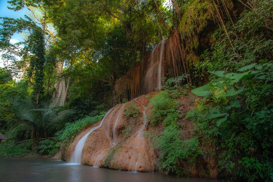 A waterfall cascades down from a limestone cliff, with a river in the forest in front, bathed in sunlight.