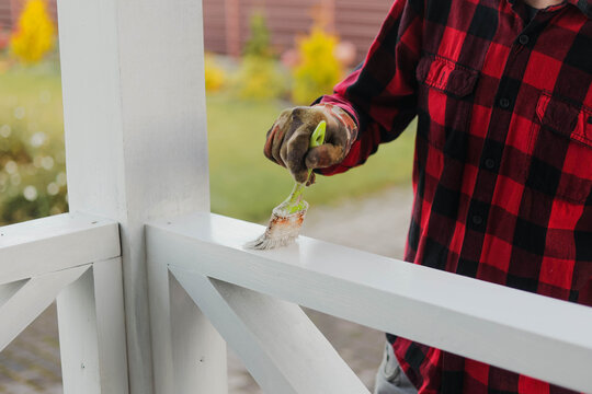 Homeowner painting a white wooden porch railing with a brush, performing exterior renovation and maintenance work outdoors.