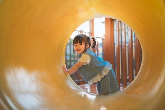 Happy toddler girl exploring colorful playground tunnel outdoors, smiling and looking back while holding the entrance of play equipment, enjoying childhood adventure, curiosity