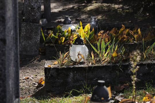 Cemetery graves with glass memorial candles. Autumn plants around stone tombs. Traditional remembrance lanterns. Graveyard background. Seasonal burial place. Quiet memorial setting.