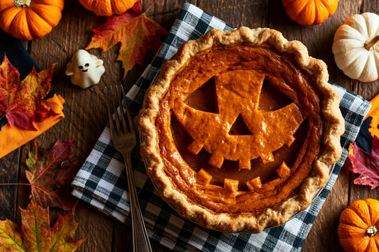 Pumpkin pie with jack-o-lantern face cut into crust, Halloween overhead flat lay with pumpkins and gingham napkin