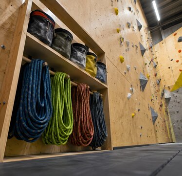 Climbing gym shelf holding chalk bags and coiled dynamic ropes against a plywood wall covered with bolts and matting