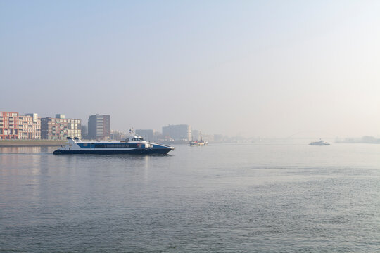 A passenger fast ferry approaches a busy river terminal in soft morning haze during rush hour. Ideal for travel, transportation, tourism, and waterfront infrastructure concepts.