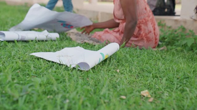 Woman in pink dress sits on grass unrolling white papers for art session. Another woman legs are visible nearby as scene takes place outdoors outdoor family