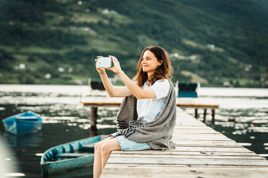 Profile view of young woman wrapped in blanket using smartphone on wooden pier by mountain lake.